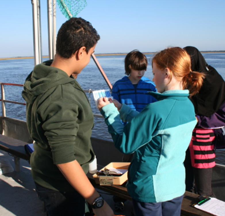 A group of thee students from a local school are huddled around one girl who is holding a water quality strip and chart.